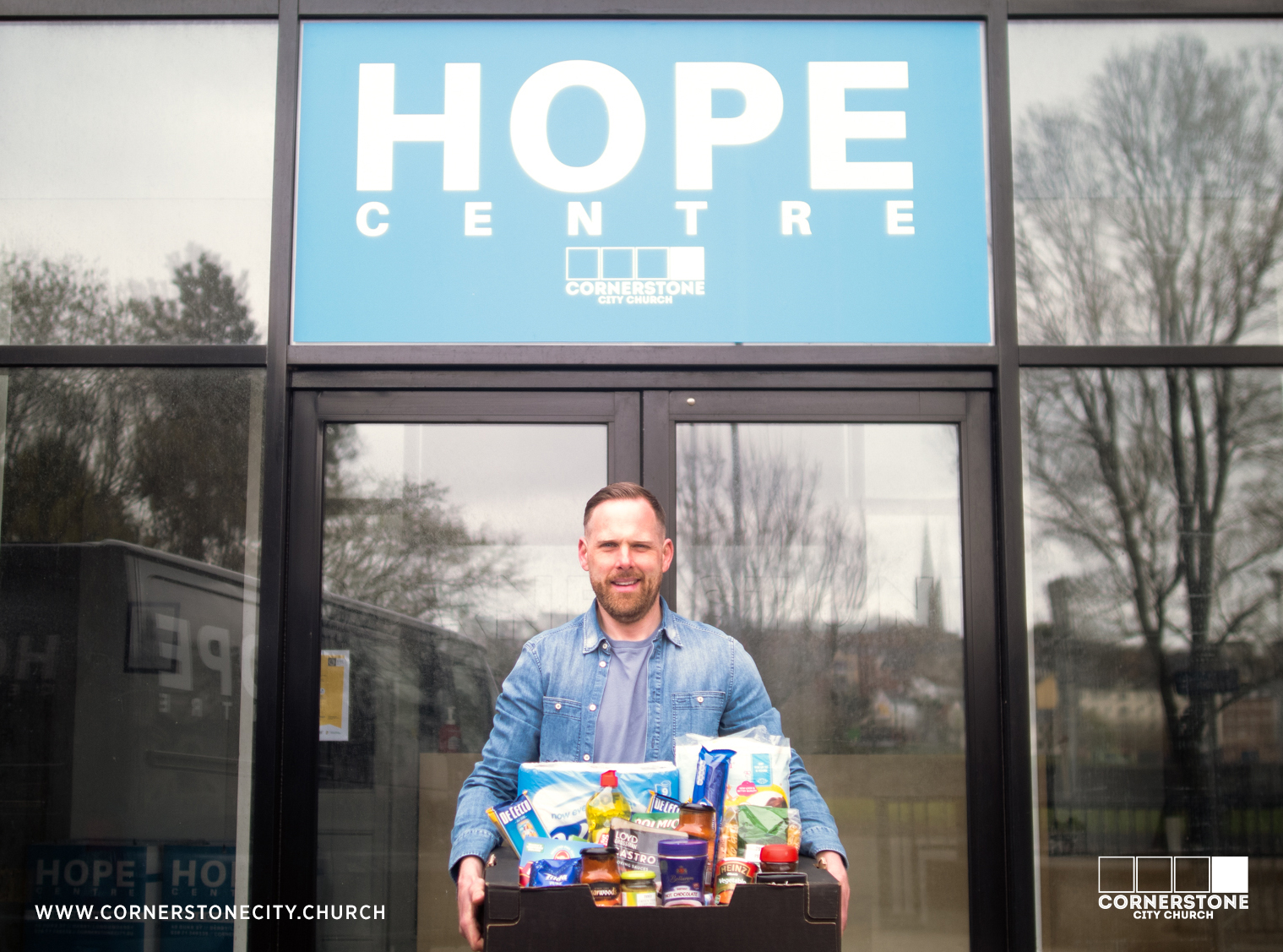 Man in a denim shirt holding a box of food to be donated to someone, outside a bulding that has a sign with the words Hope Centre