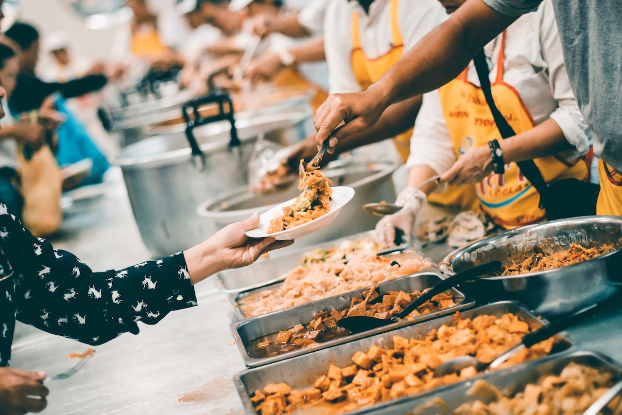 A row of volunteers scooping out food from trays to people with their hands stretched out.