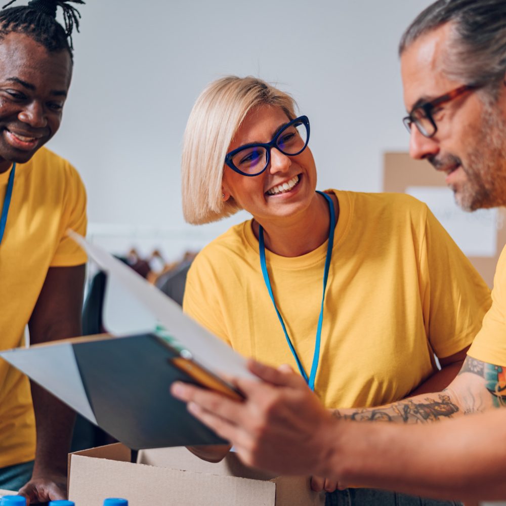 Group of diverse people working in charitable foundation. Happy volunteer separating donation stuff. Social workers making check list of the things that are done. Working as a team.